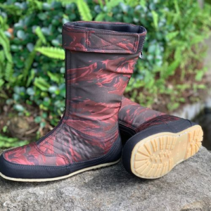 The image shows a pair of tall boots with a reddish-brown camouflage pattern. The boots have a black fabric upper and a cream-colored sole. They are shown outdoors, with lush green foliage in the background.