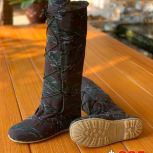 The photo shows a pair of tall boots with a camouflage pattern. The boots are laced up the front and have a flat sole. They are sitting on a wooden surface. The camouflage pattern appears to be a tiger stripe design in muted greens and browns.