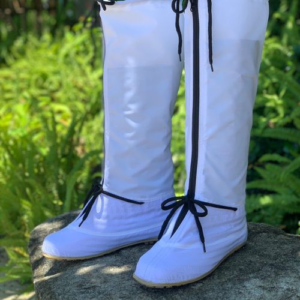 The image shows a pair of white, tall, waterproof boots. They have black laces and zippers. The boots are shown outdoors, on a rock, with green foliage in the background.