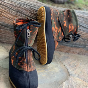 The image shows a pair of ankle-high boots. The boots are predominantly black with an orange and green camouflage pattern on the upper part. They have laces and zippers for closure, and the soles appear to be made of a durable material. The boots are shown on a rocky surface.
