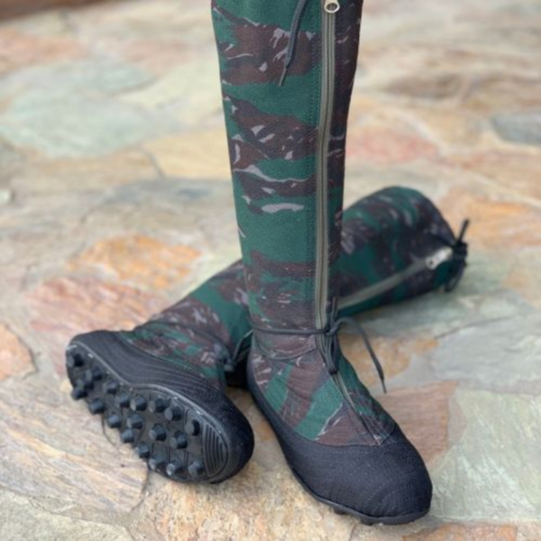 The image shows a pair of tall, camouflage-patterned boots. The boots are predominantly dark green and brown, with a camouflage pattern. They have black soles with small studs and appear to be made of a fabric material. A zipper is visible on the side of each boot. The boots are positioned on a stone or paved surface.